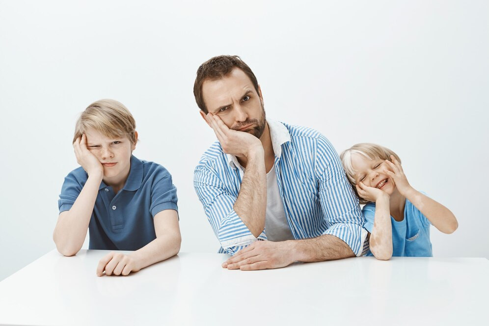 Retrato familia divertida padre e hijos sentados mesa inclinando cabeza mano haciendo muecas 176420 24797