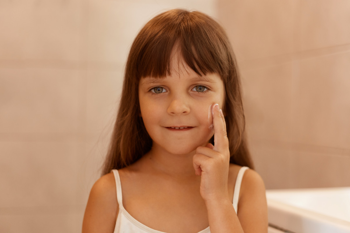 Toma en interiores de una nina con crema facial en la mejilla senalando con el dedo en la cara mirando directamente la camara vistiendo una camiseta blanca sin mangas realizando procedimientos de belleza sola